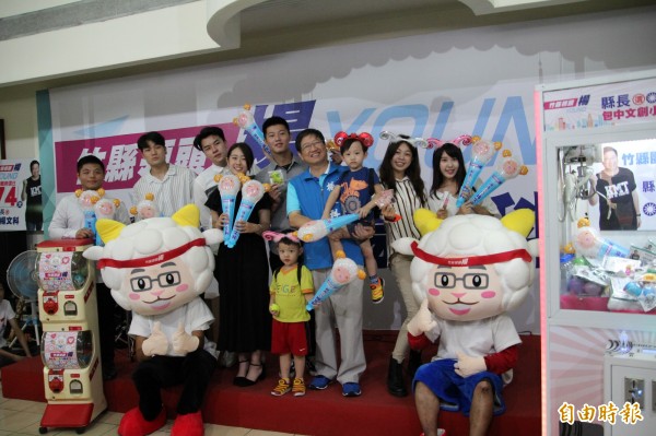 國民黨新竹縣長參選人楊文科(著藍背心者)今天發表他的競選主題曲及多項「包中文創小物」,邀請大家一起參加他的「藝起來飛揚」活動,玩「領頭楊」的夾娃娃機(圖右)或扭蛋(圖左下)就可以免費獲得。(記者黃美珠攝)