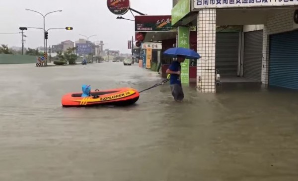 1名父親撐著雨傘，拉著1條充氣小船，船中坐著1名穿著藍色牛角造型雨衣的女童。（圖擷取自爆廢公社）