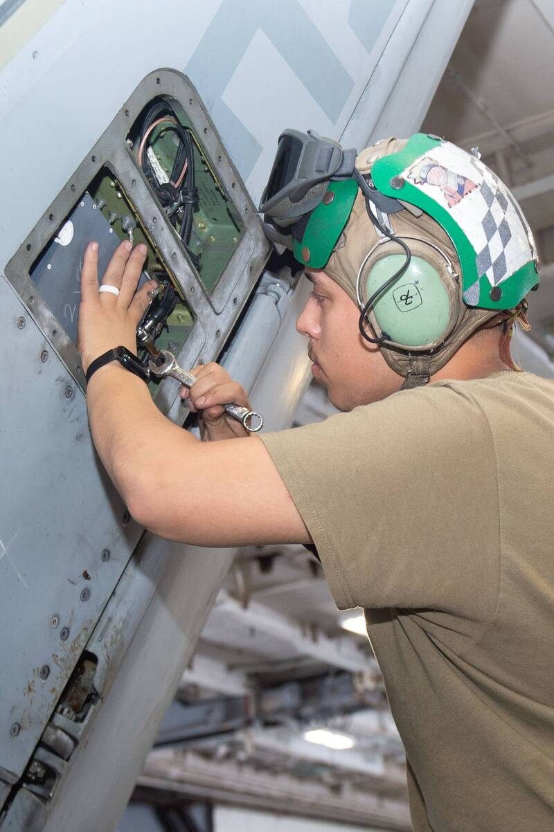 地勤保修官兵於機庫內針對F-18戰機的垂直尾翼進行檢修，圖中可見機身的蒙皮艙蓋已被打開，露出部分的內部線路結構。（圖片擷取自第七艦隊臉書）