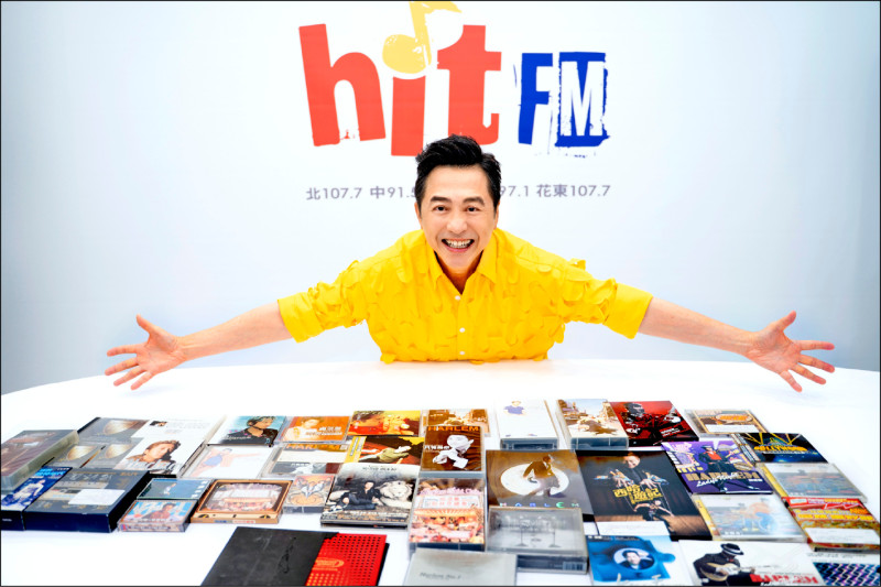 庾澄慶がHit FMのスタジオで、机に並べた自身の作品を前に両手を広げている