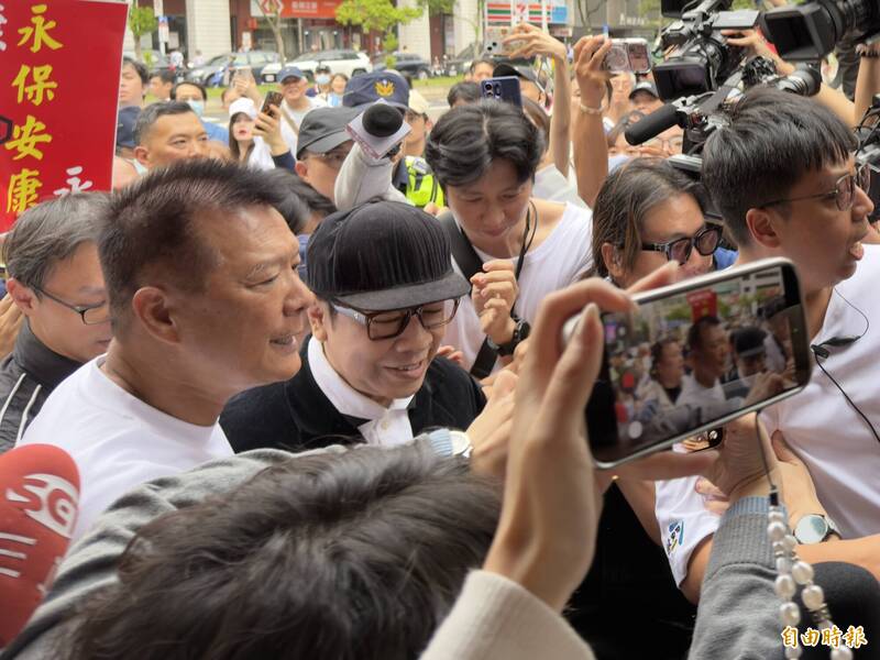 蔡康永戴着黑帽、穿黑色西装,在蔣万安竞选总部成立活动现场被媒体与人群围住