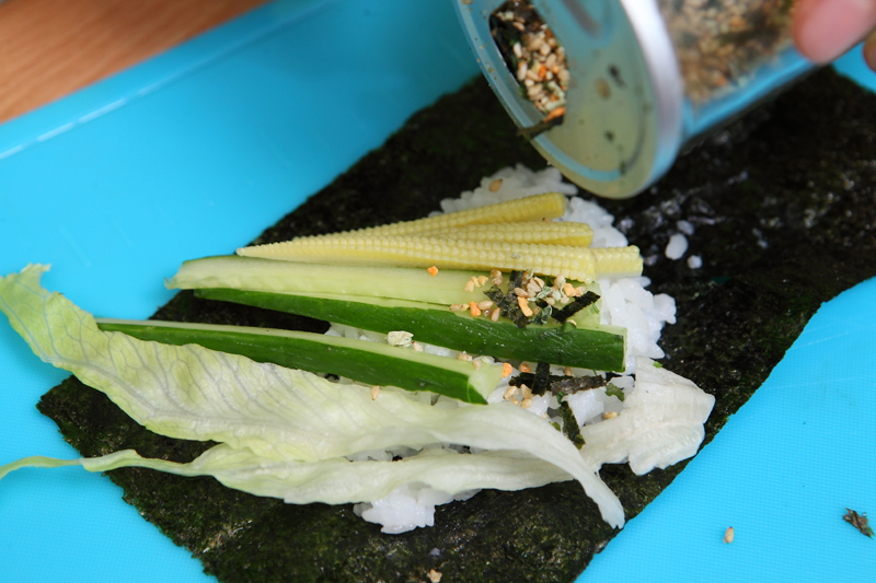 取壽司海苔斜放上醋飯、小黃瓜段、玉米筍、萵苣葉絲，再撒上適量香鬆。