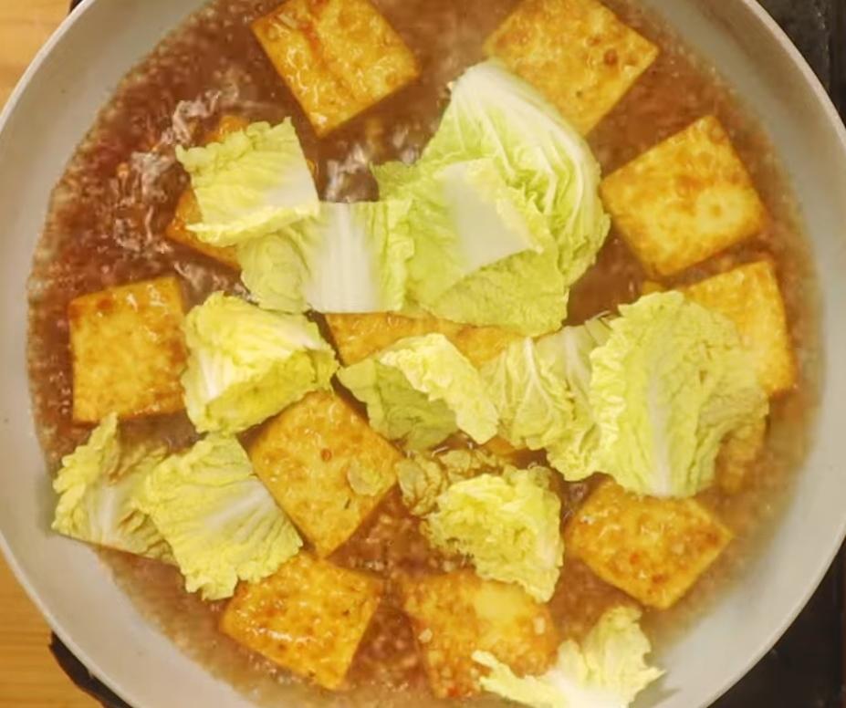 豆腐推至鍋邊,放入蒜仁、嫩薑炒香,再下辣豆瓣醬跟醬油炒出香氣,接著加入適量水、娃娃菜、糖、白胡椒跟醬色煨煮。