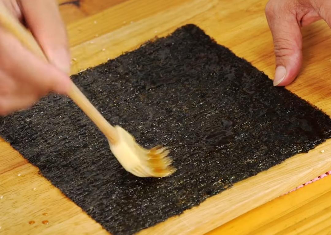 取一張海苔刷上煮好的醬汁,均勻撒上白芝麻,取另一張海苔片一樣刷上醬汁,將其蓋上,使用擀麵棍將其擀至黏合,平放在烤網上。