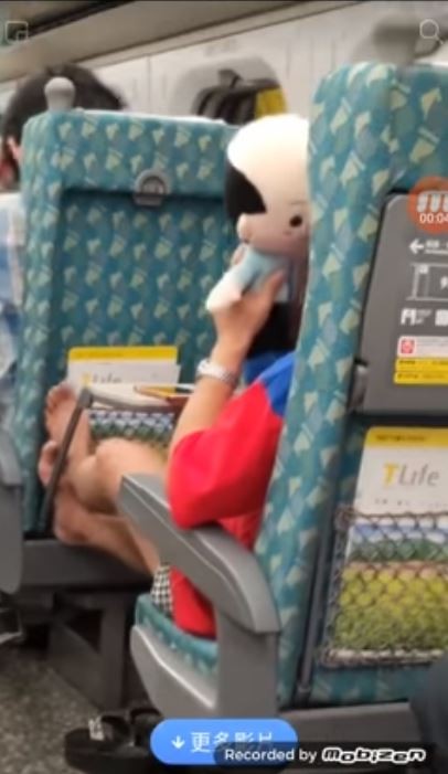 一名疑似韓粉在高鐵上把腳翹上前方的椅背，一邊親韓國瑜造型玩偶，一邊「搓腳」，引起討論。（擷取自「爆怨公社」影片）