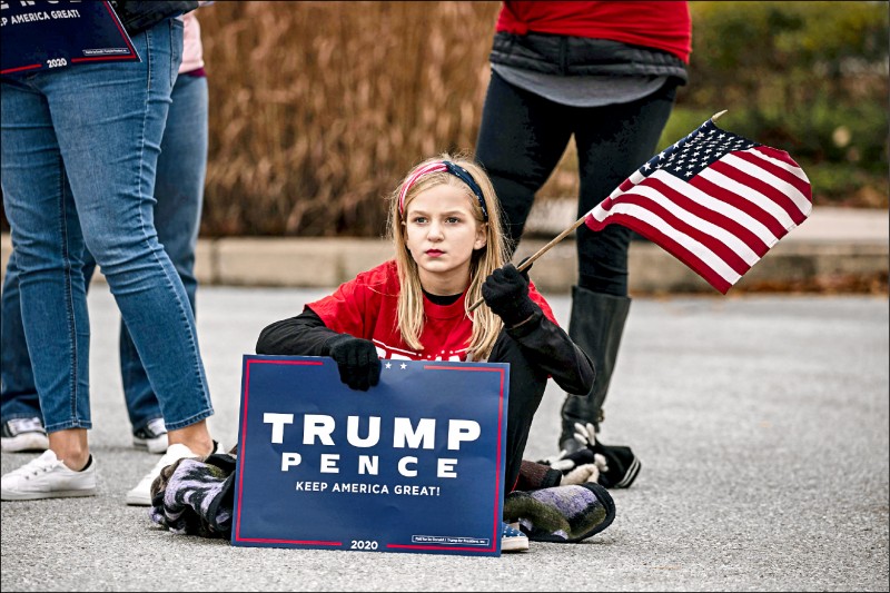 美國賓州參議院多數政策委員會廿五日舉行大選聽證會的溫德姆蓋茲堡飯店外，一名支持川普的女孩坐在地上，並揮舞美國國旗。（法新社）