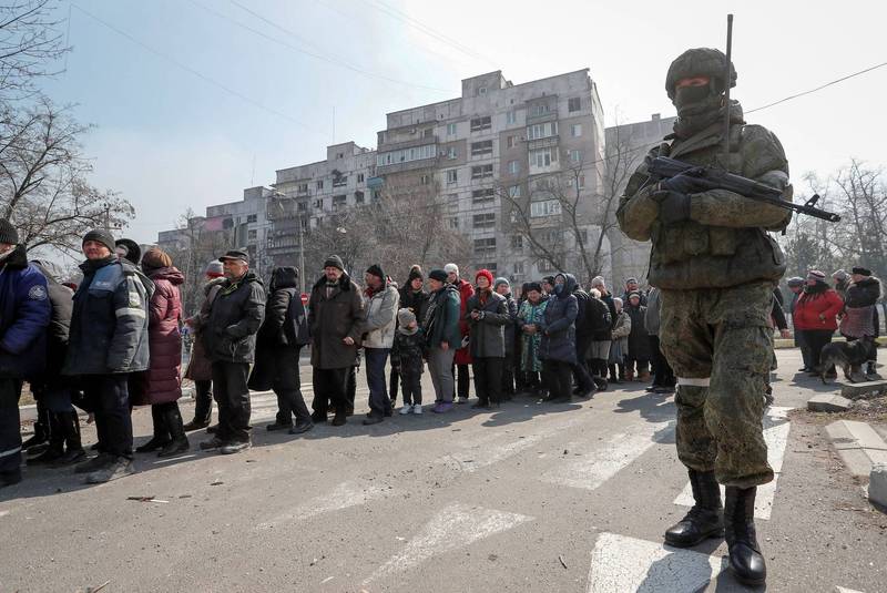 烏克蘭馬利波市，一名俄軍士兵持槍警戒。烏克蘭民眾則在他身後排隊領取人道救援物資。（路透）
