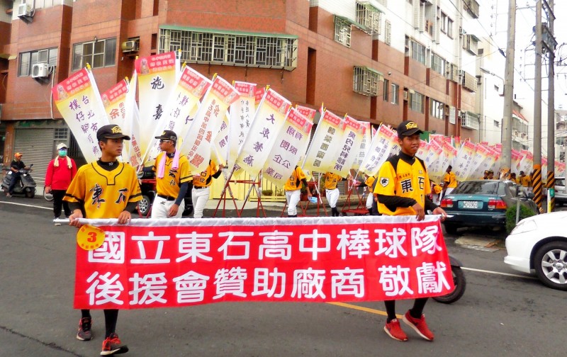 東石高中棒球隊以往參加配天宮媽祖誕辰遶境慶典，會扛著印著贊助商名稱的大旗遊行。（記者林宜樟翻攝）