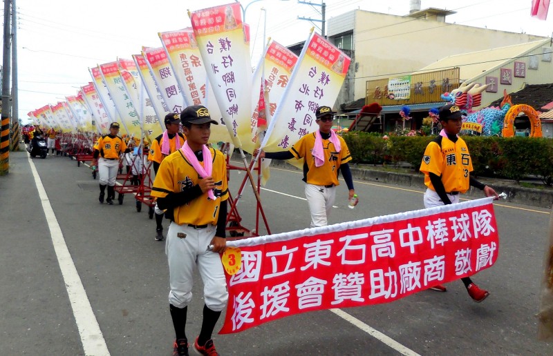 東石高中棒球隊以往參加配天宮媽祖誕辰遶境慶典，會扛著印著贊助商名稱的大旗遊行。（記者林宜樟翻攝）