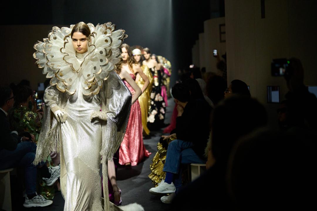 香檳金禮服搭配層層堆疊的誇張立領設計，透過光澤面料與雕塑輪廓，強化角色性與舞台張力，呼應系列對「凝視」的主題思考。