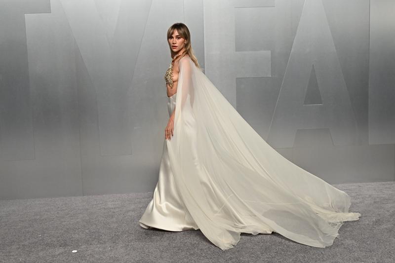 Suki Waterhouse一襲象牙白緞面禮服搭配飄逸披風，頸部裝飾立體金色飾件，整體風格充滿古典戲劇感。